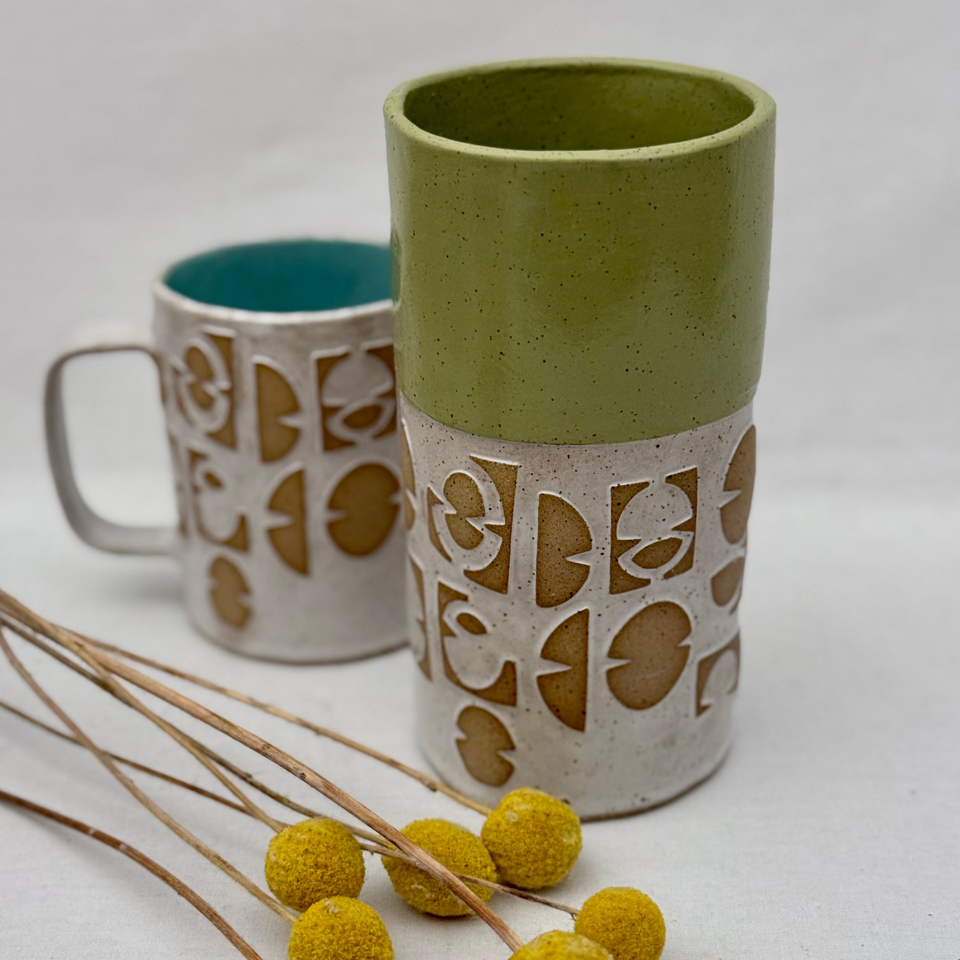 ceramic vase with geometric patterns with matching mug behind it and a small arrangement of yellow flowers on a white background