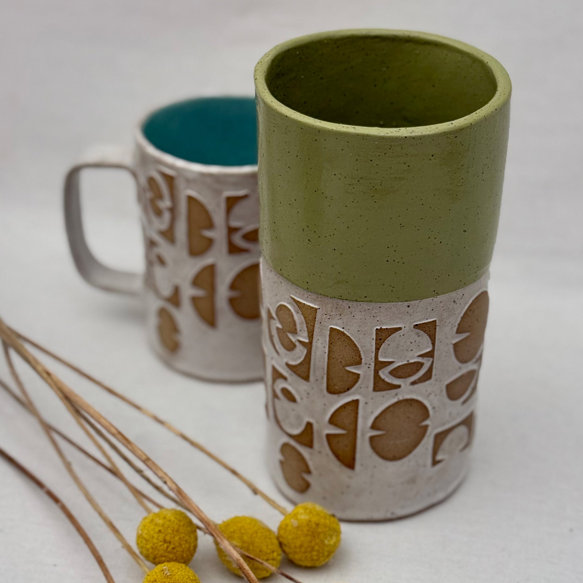 ceramic vase with geometric patterns with matching mug behind it and a small arrangement of yellow flowers on a white background