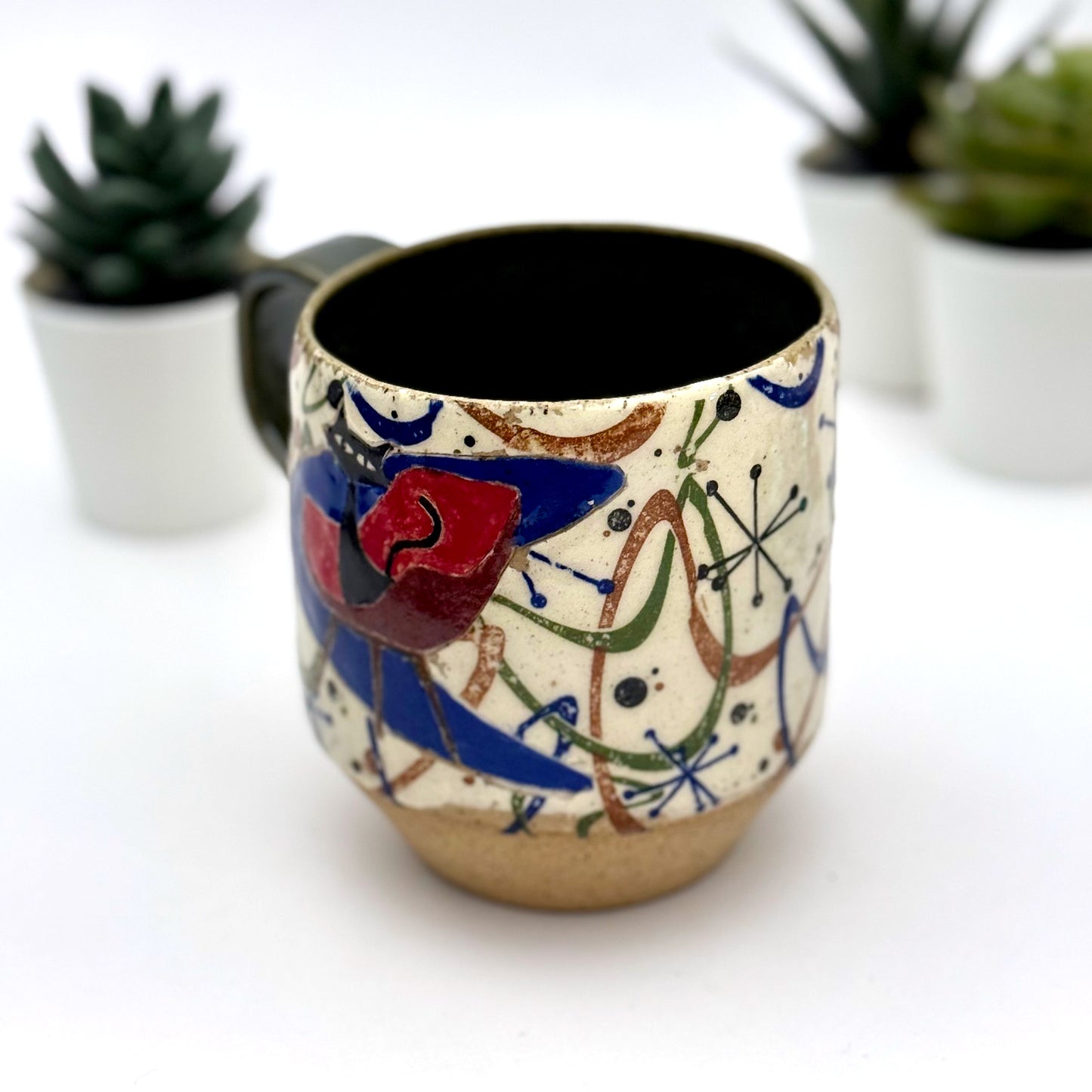 A retro-inspired ceramic mug featuring boomerang motifs, a lounging black cat in a chair, and a large cat face graphic on the opposite side, shown with a dark interior glaze.