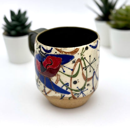 A retro-inspired ceramic mug featuring boomerang motifs, a lounging black cat in a chair, and a large cat face graphic on the opposite side, shown with a dark interior glaze.