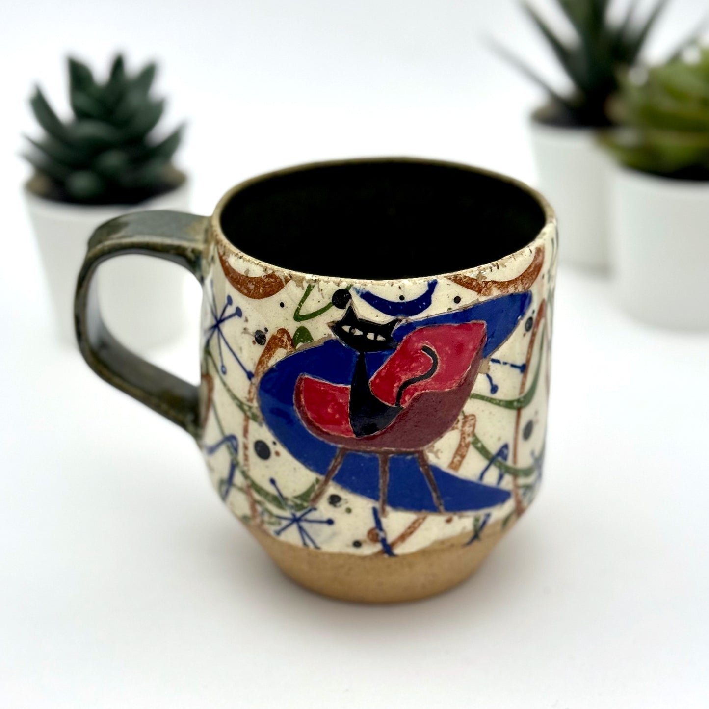 A retro-inspired ceramic mug featuring boomerang motifs, a lounging black cat in a chair, and a large cat face graphic on the opposite side, shown with a dark interior glaze.