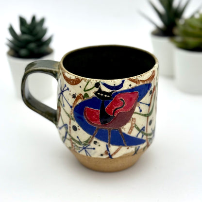 A retro-inspired ceramic mug featuring boomerang motifs, a lounging black cat in a chair, and a large cat face graphic on the opposite side, shown with a dark interior glaze.