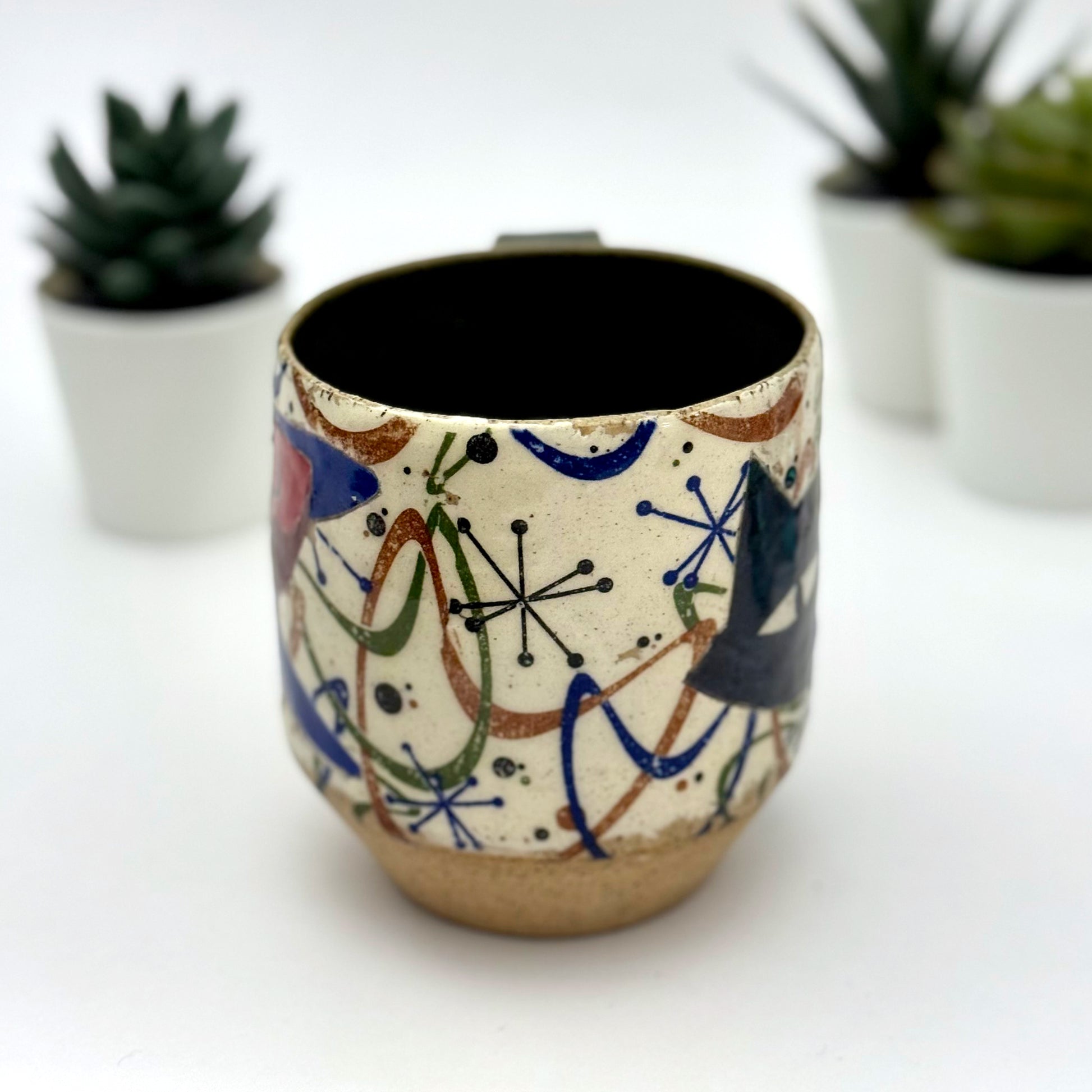 A retro-inspired ceramic mug featuring boomerang motifs, a lounging black cat in a chair, and a large cat face graphic on the opposite side, shown with a dark interior glaze.