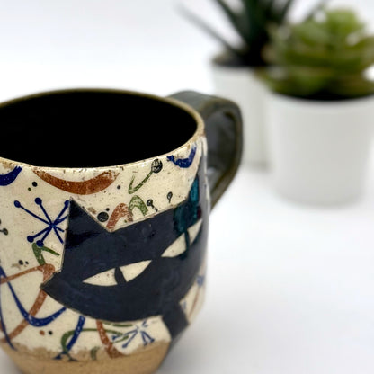 A retro-inspired ceramic mug featuring boomerang motifs, a lounging black cat in a chair, and a large cat face graphic on the opposite side, shown with a dark interior glaze.