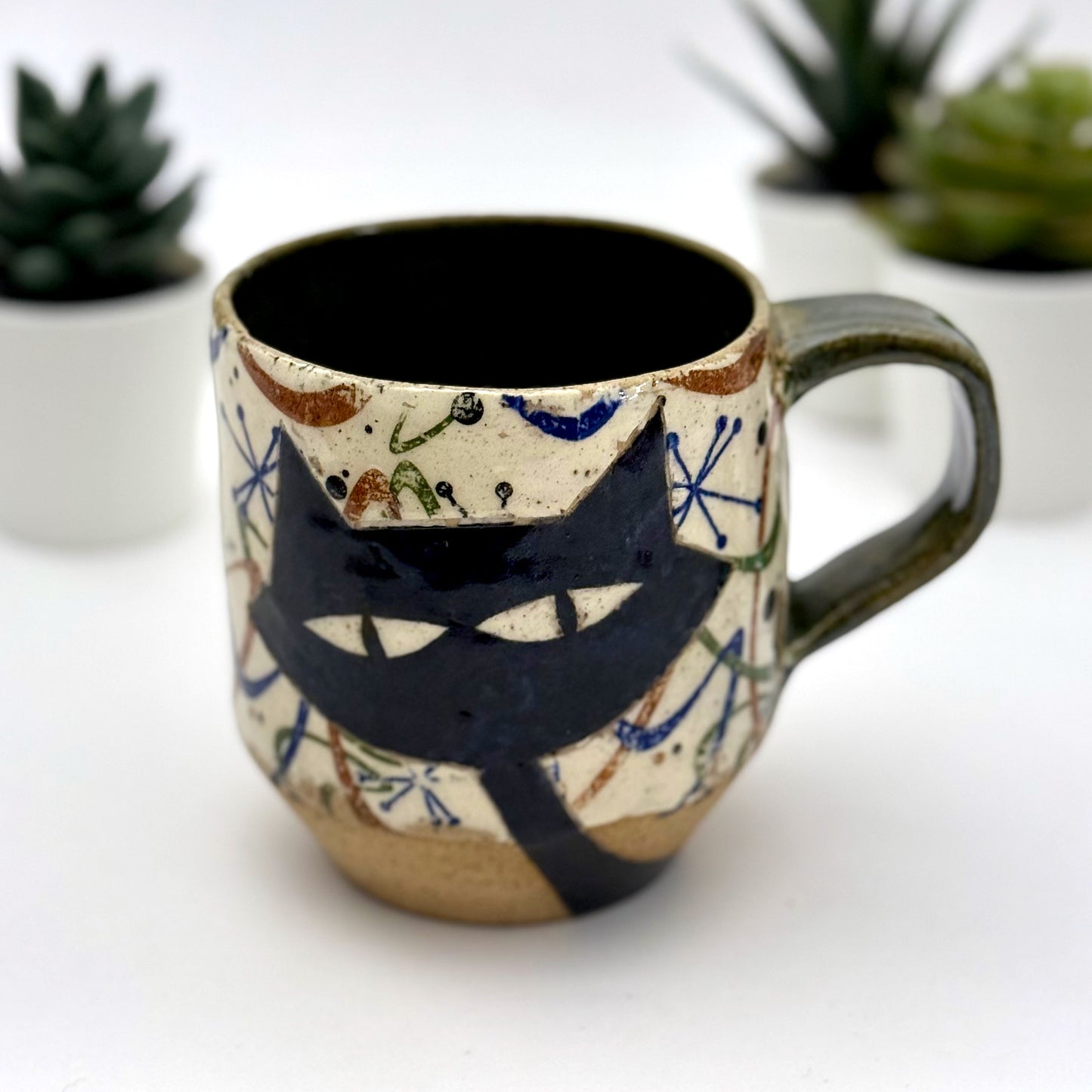 A retro-inspired ceramic mug featuring boomerang motifs, a lounging black cat in a chair, and a large cat face graphic on the opposite side, shown with a dark interior glaze.