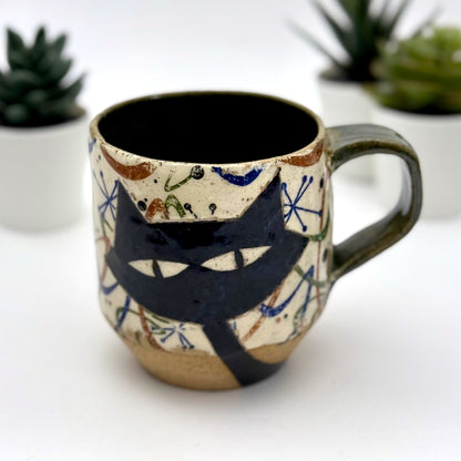 A retro-inspired ceramic mug featuring boomerang motifs, a lounging black cat in a chair, and a large cat face graphic on the opposite side, shown with a dark interior glaze.
