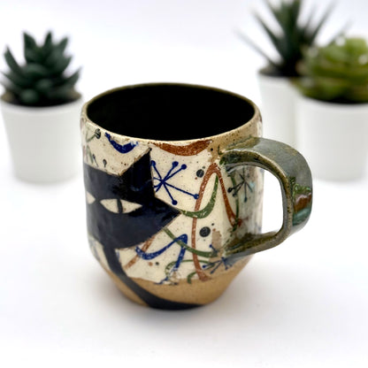 A retro-inspired ceramic mug featuring boomerang motifs, a lounging black cat in a chair, and a large cat face graphic on the opposite side, shown with a dark interior glaze.