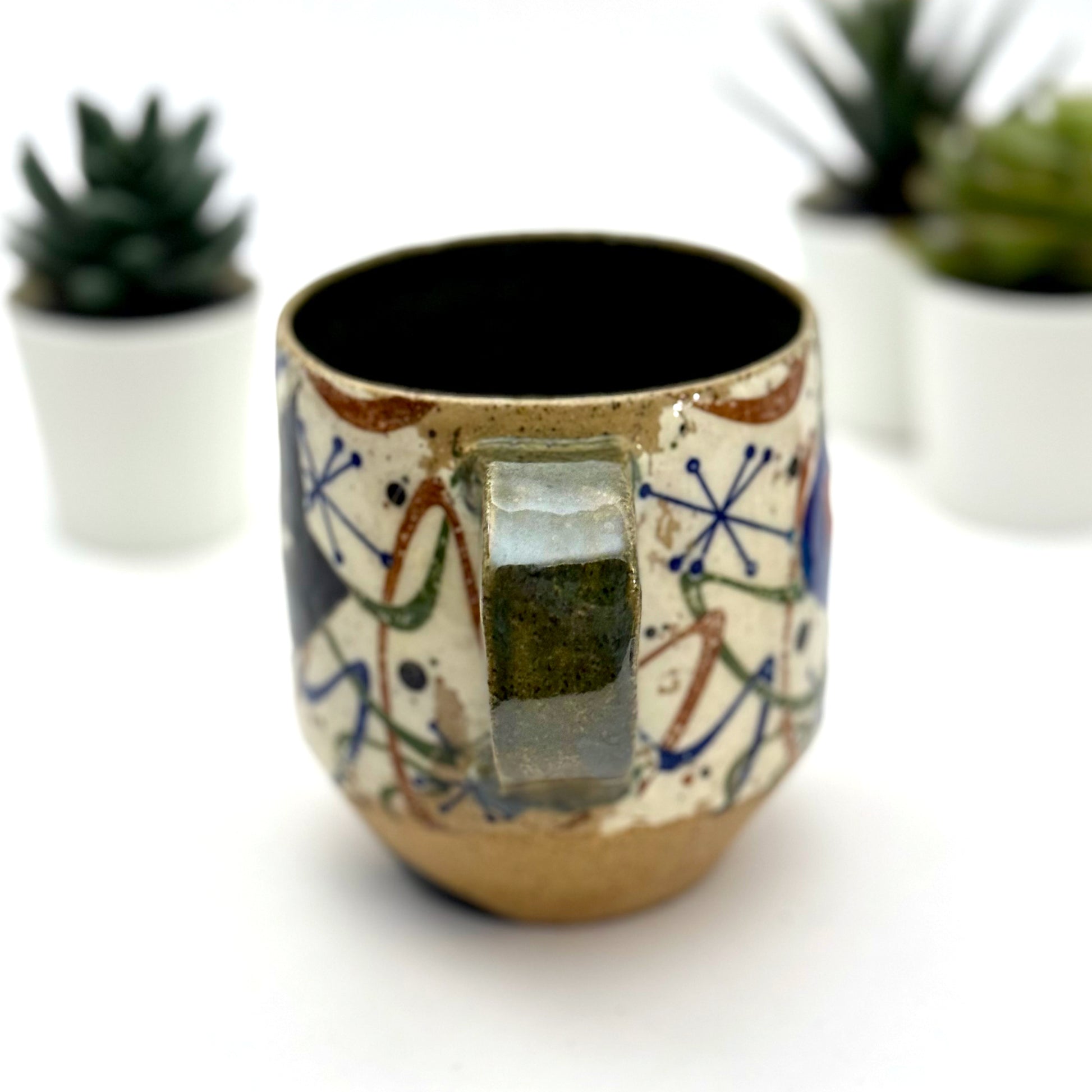 A retro-inspired ceramic mug featuring boomerang motifs, a lounging black cat in a chair, and a large cat face graphic on the opposite side, shown with a dark interior glaze.