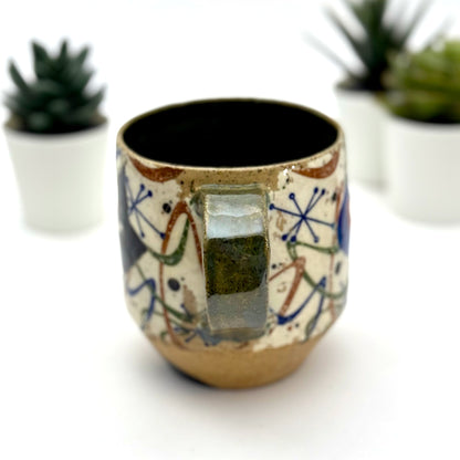 A retro-inspired ceramic mug featuring boomerang motifs, a lounging black cat in a chair, and a large cat face graphic on the opposite side, shown with a dark interior glaze.