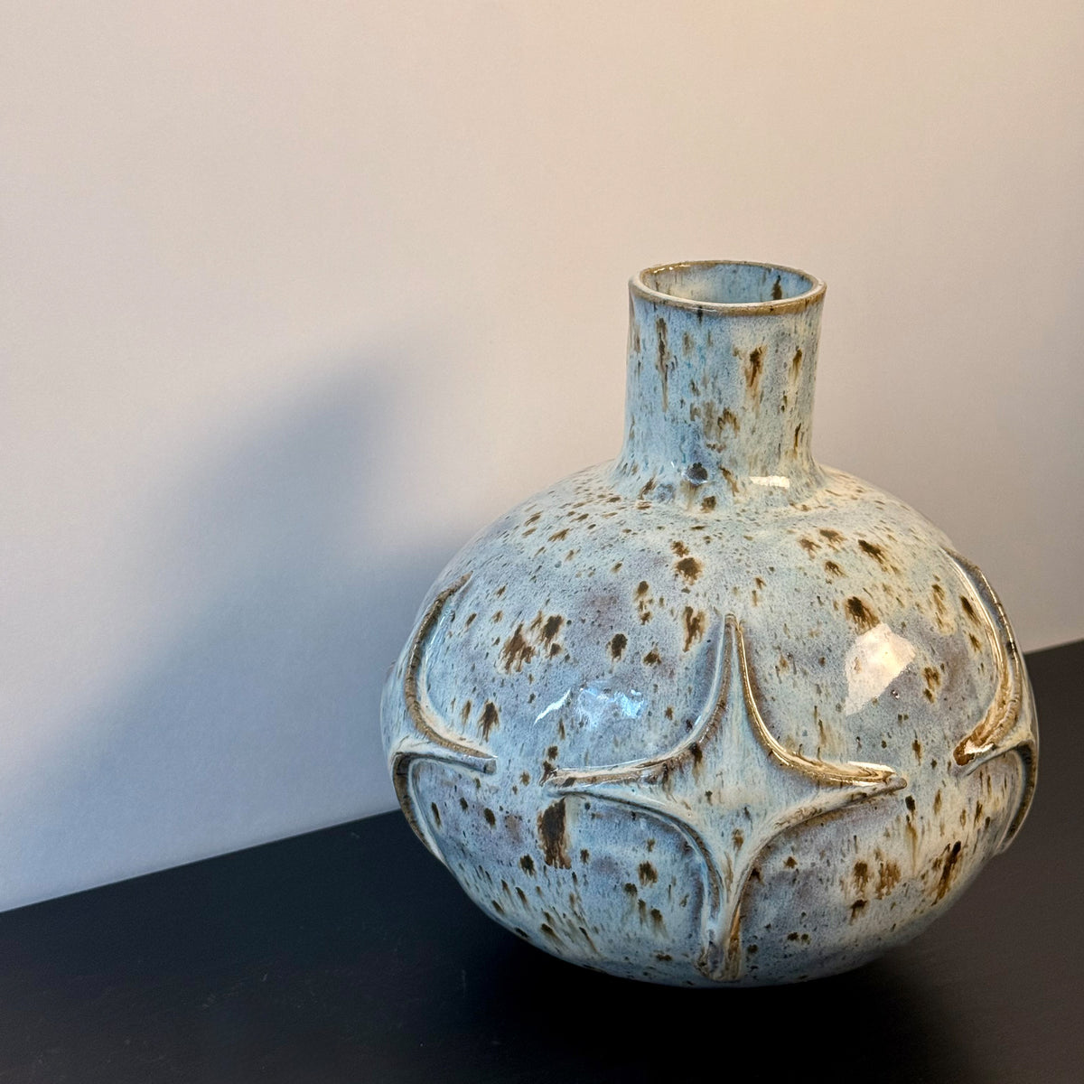 Handmade ceramic vase with a rounded sculptural body, narrow cylindrical neck, and raised atomic starburst motifs around the lower half. Finished in a pale blue-and-cream mottled glaze with darker speckled accents that highlight the carved geometric details.