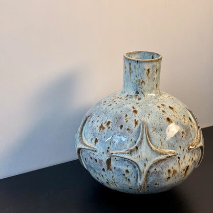 Handmade ceramic vase with a rounded sculptural body, narrow cylindrical neck, and raised atomic starburst motifs around the lower half. Finished in a pale blue-and-cream mottled glaze with darker speckled accents that highlight the carved geometric details.
