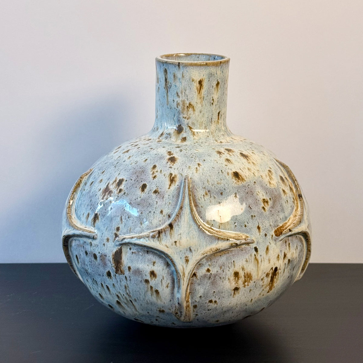 Handmade ceramic vase with a rounded sculptural body, narrow cylindrical neck, and raised atomic starburst motifs around the lower half. Finished in a pale blue-and-cream mottled glaze with darker speckled accents that highlight the carved geometric details.