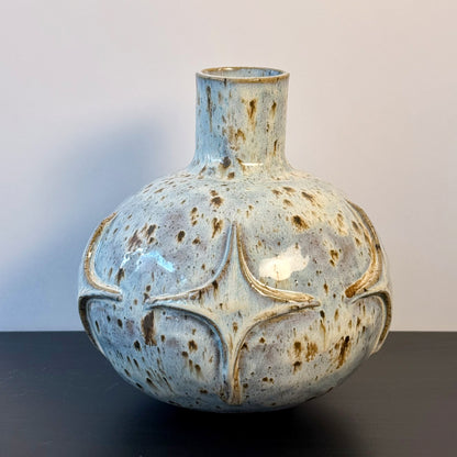 Handmade ceramic vase with a rounded sculptural body, narrow cylindrical neck, and raised atomic starburst motifs around the lower half. Finished in a pale blue-and-cream mottled glaze with darker speckled accents that highlight the carved geometric details.