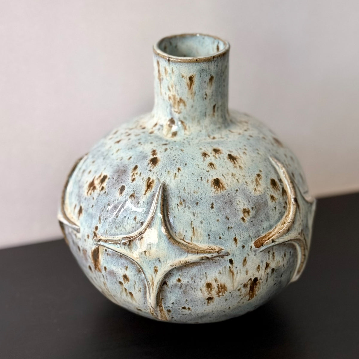 Handmade ceramic vase with a rounded sculptural body, narrow cylindrical neck, and raised atomic starburst motifs around the lower half. Finished in a pale blue-and-cream mottled glaze with darker speckled accents that highlight the carved geometric details.