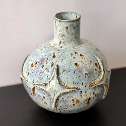 Handmade ceramic vase with a rounded sculptural body, narrow cylindrical neck, and raised atomic starburst motifs around the lower half. Finished in a pale blue-and-cream mottled glaze with darker speckled accents that highlight the carved geometric details.
