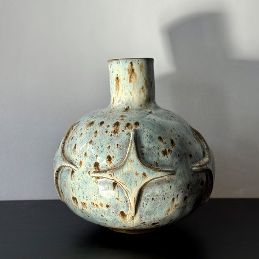 Handmade ceramic vase with a rounded sculptural body, narrow cylindrical neck, and raised atomic starburst motifs around the lower half. Finished in a pale blue-and-cream mottled glaze with darker speckled accents that highlight the carved geometric details.