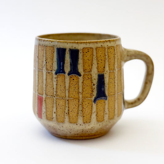 Hand-thrown stoneware mug with a warm speckled glaze and retro geometric grid pattern in tan, deep blue, gray-blue, and muted coral accents. The rounded mug features a curved handle, glossy finish, and mid-century-inspired design. Photographed against a soft white background.