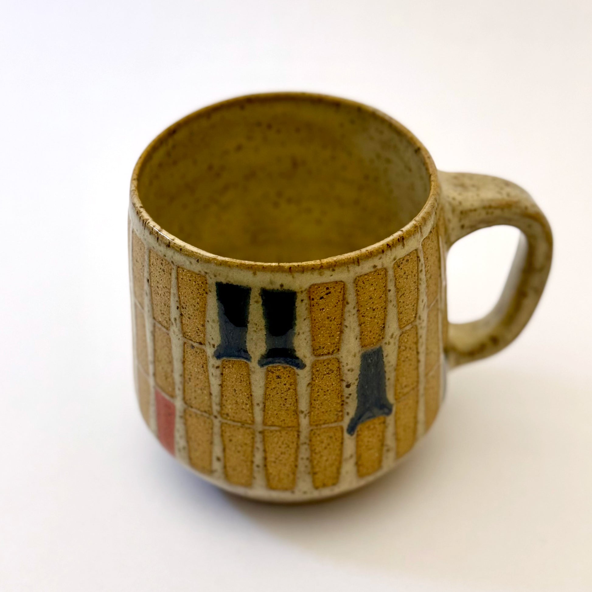 Hand-thrown stoneware mug with a warm speckled glaze and retro geometric grid pattern in tan, deep blue, gray-blue, and muted coral accents. The rounded mug features a curved handle, glossy finish, and mid-century-inspired design. Photographed against a soft white background.