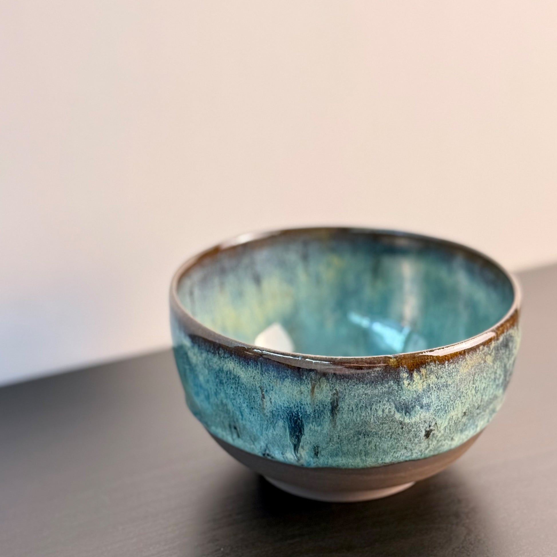 A handmade ceramic bowl with a glossy, mottled glaze of teal, blue, and brown, sitting on a dark wooden surface against a plain light background.