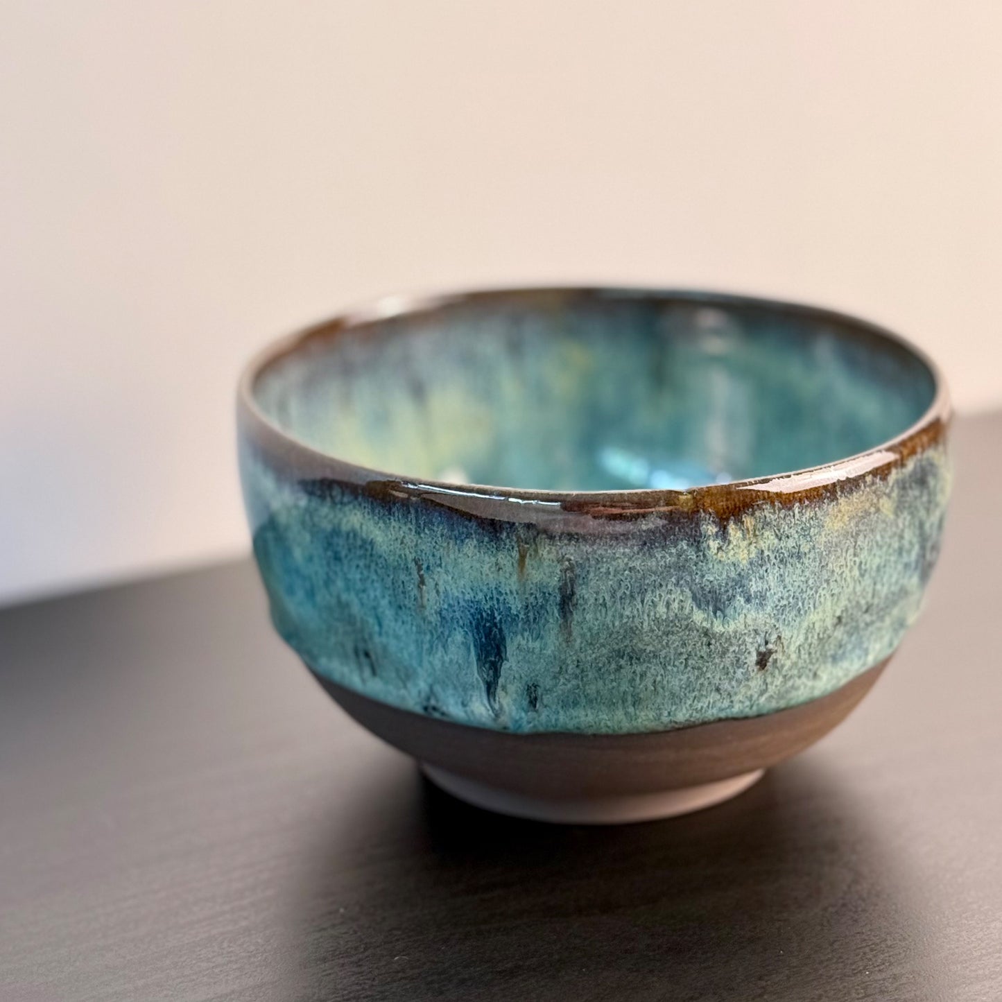 A handmade ceramic bowl with a textured, mottled blue and green glaze and a natural brown clay base, shown on a dark wooden surface against a neutral beige background.