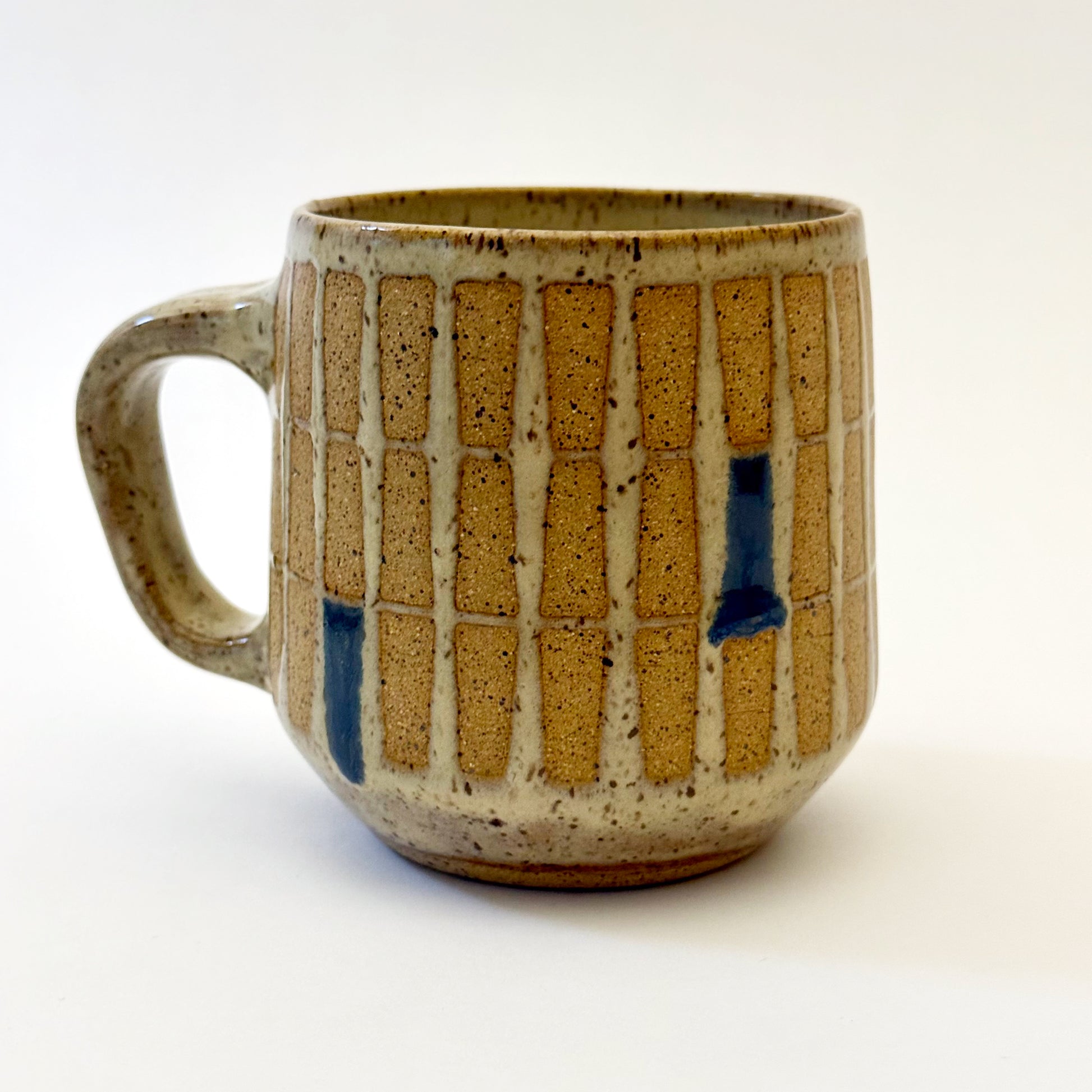 Hand-thrown stoneware mug with a warm speckled glaze and retro geometric grid pattern in tan, deep blue, gray-blue, and muted coral accents. The rounded mug features a curved handle, glossy finish, and mid-century-inspired design. Photographed against a soft white background.