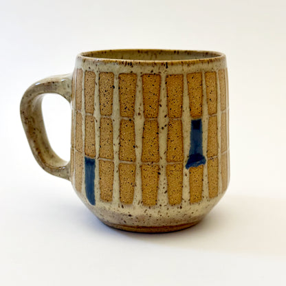Hand-thrown stoneware mug with a warm speckled glaze and retro geometric grid pattern in tan, deep blue, gray-blue, and muted coral accents. The rounded mug features a curved handle, glossy finish, and mid-century-inspired design. Photographed against a soft white background.
