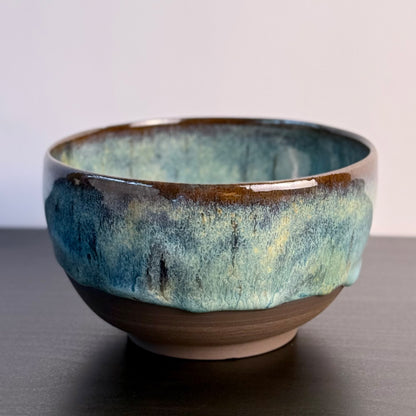 A handmade ceramic bowl with a glossy glaze featuring a gradient of teal, blue, and brown, resting on a dark surface against a light gray background.