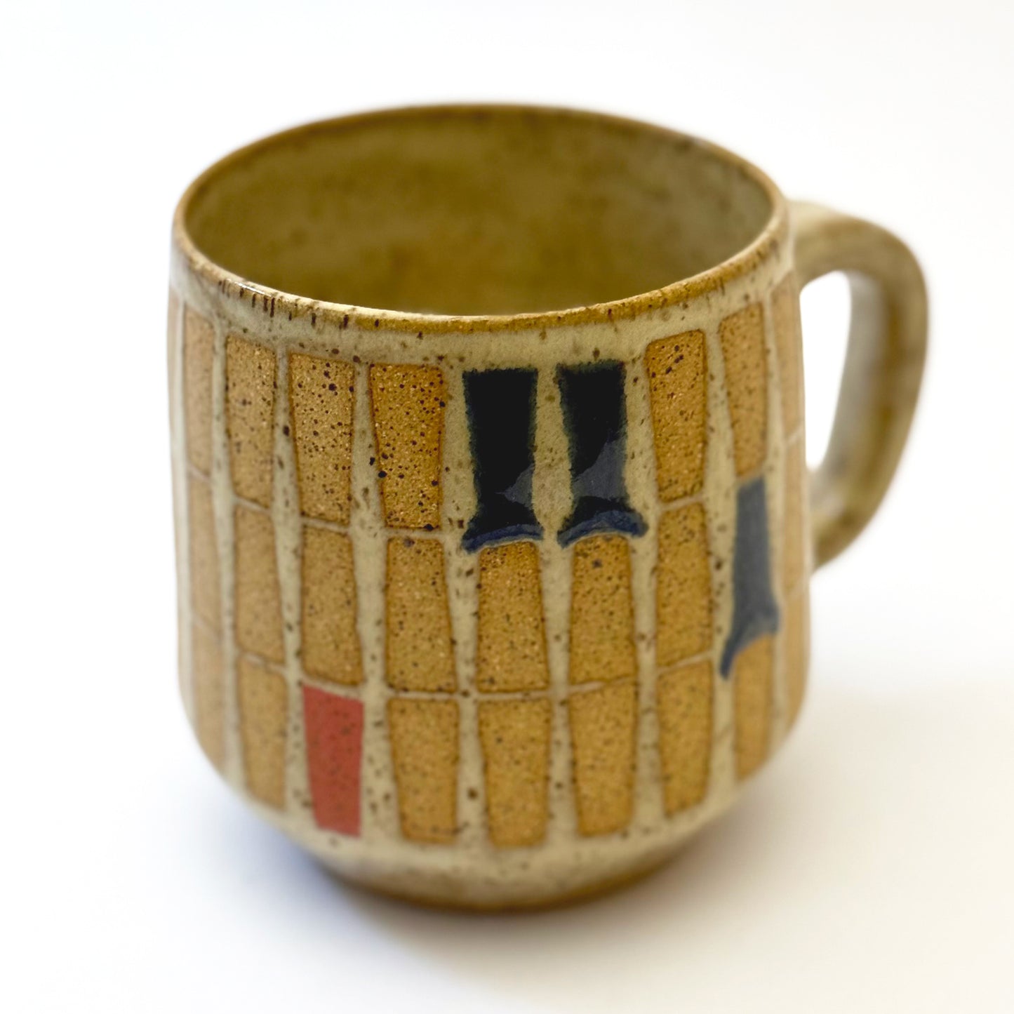 Hand-thrown stoneware mug with a warm speckled glaze and retro geometric grid pattern in tan, deep blue, gray-blue, and muted coral accents. The rounded mug features a curved handle, glossy finish, and mid-century-inspired design. Photographed against a soft white background.