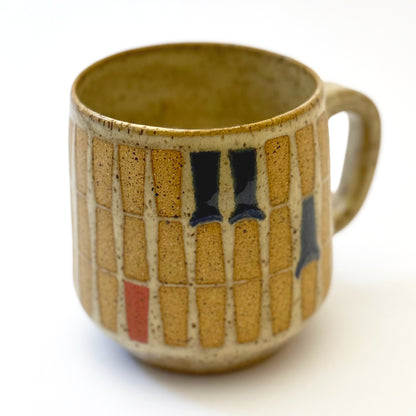 Hand-thrown stoneware mug with a warm speckled glaze and retro geometric grid pattern in tan, deep blue, gray-blue, and muted coral accents. The rounded mug features a curved handle, glossy finish, and mid-century-inspired design. Photographed against a soft white background.