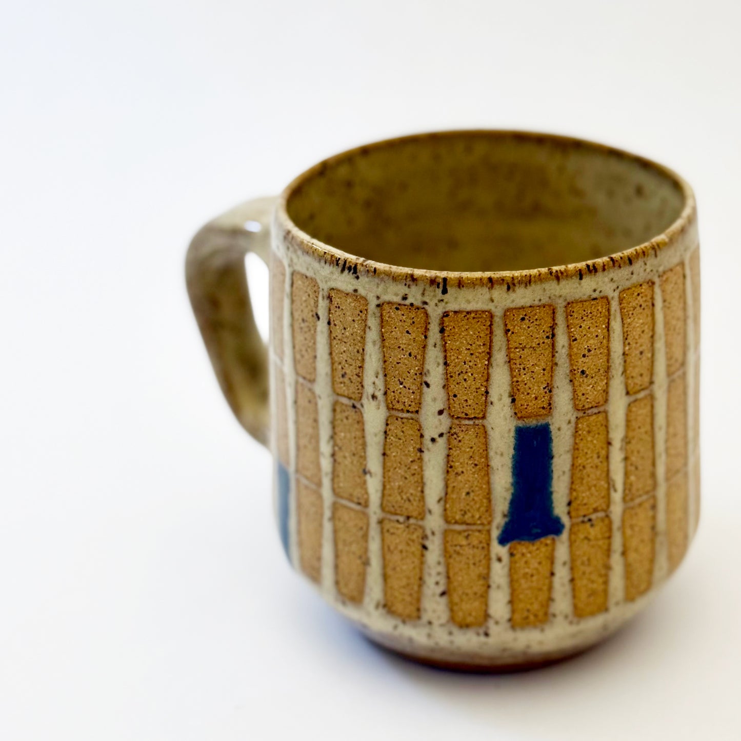 Hand-thrown stoneware mug with a warm speckled glaze and retro geometric grid pattern in tan, deep blue, gray-blue, and muted coral accents. The rounded mug features a curved handle, glossy finish, and mid-century-inspired design. Photographed against a soft white background.