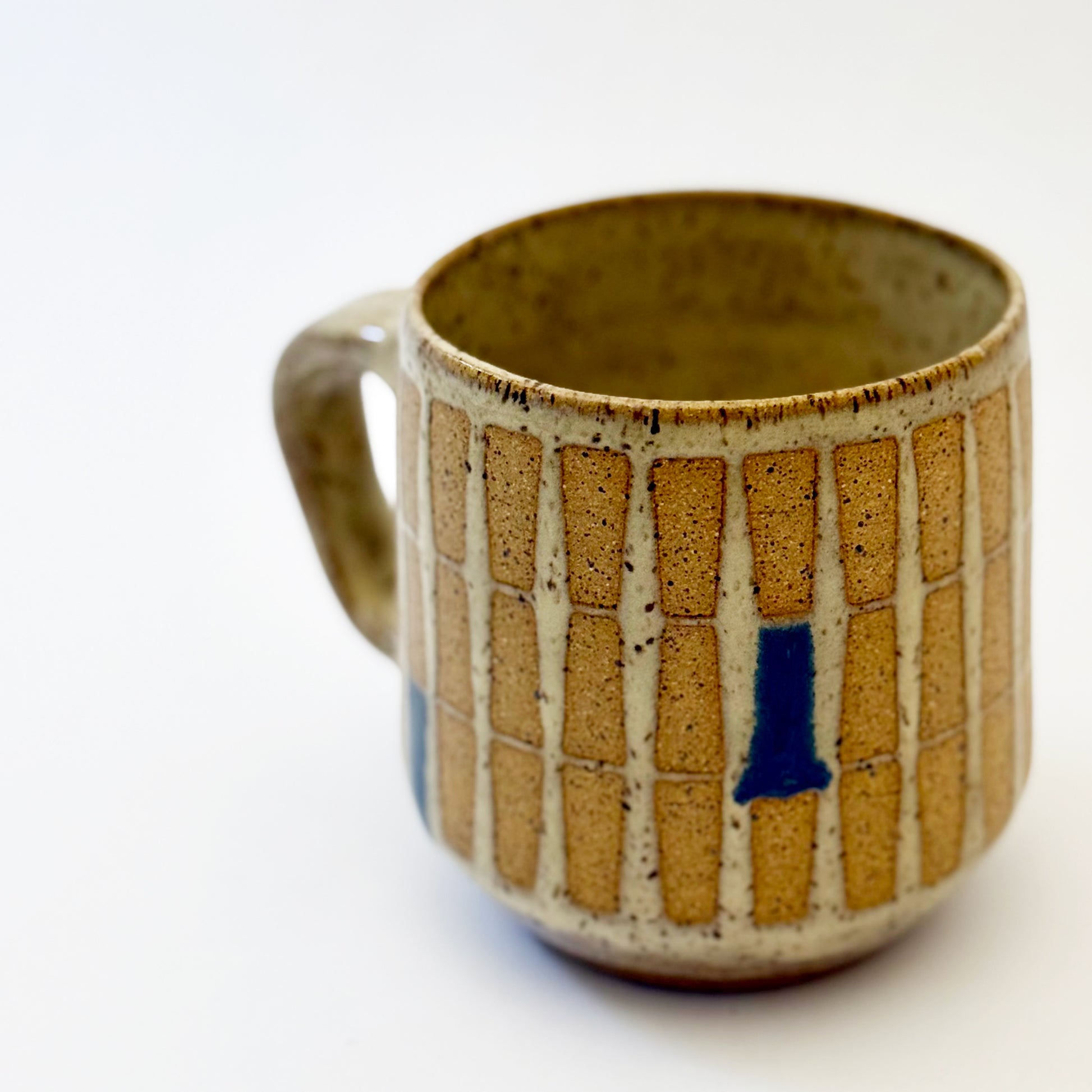 Hand-thrown stoneware mug with a warm speckled glaze and retro geometric grid pattern in tan, deep blue, gray-blue, and muted coral accents. The rounded mug features a curved handle, glossy finish, and mid-century-inspired design. Photographed against a soft white background.