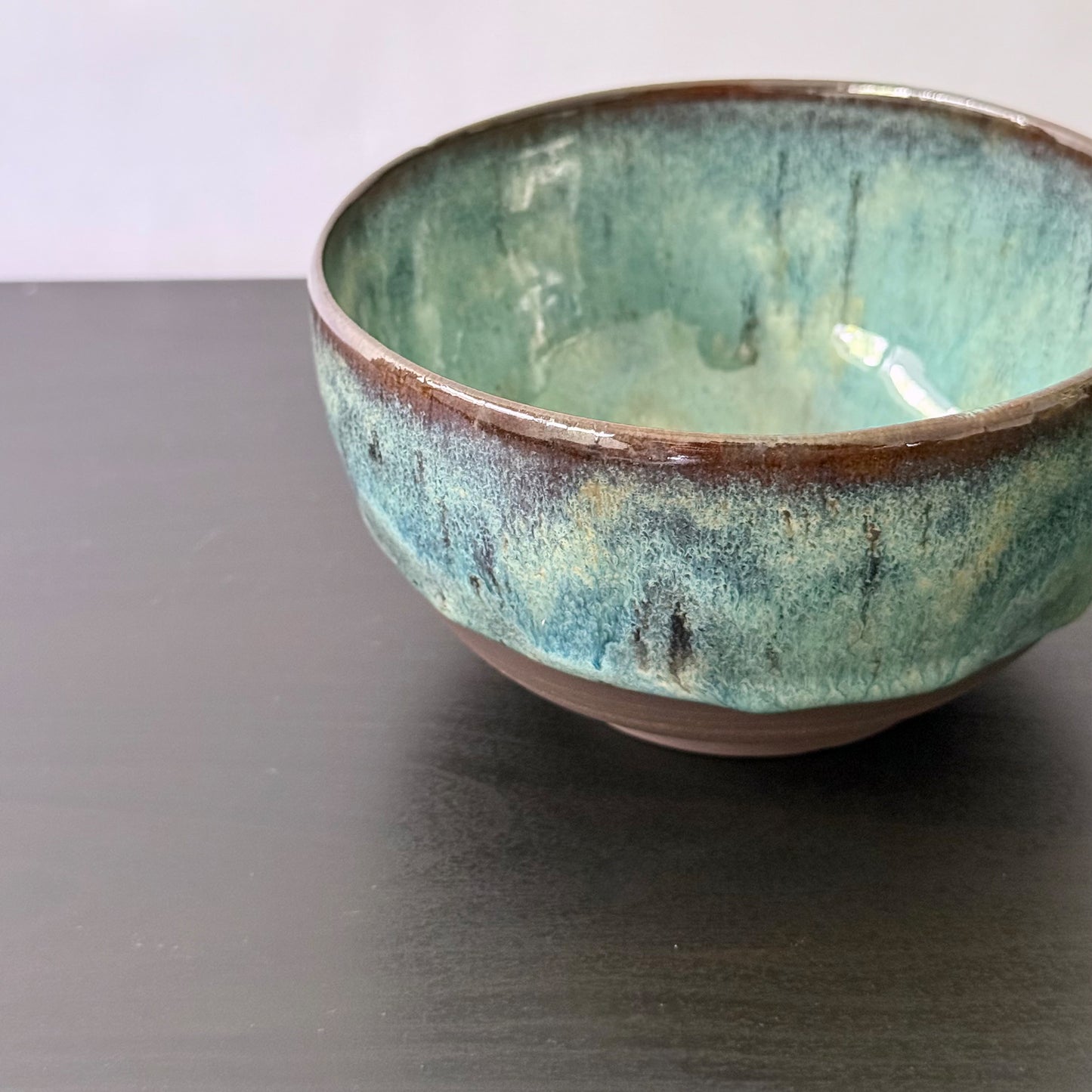 A handmade ceramic bowl with a textured glaze featuring shades of teal, green, and brown, shown on a dark tabletop.