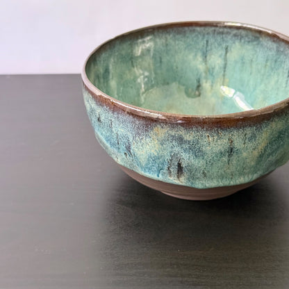 A handmade ceramic bowl with a textured glaze featuring shades of teal, green, and brown, shown on a dark tabletop.