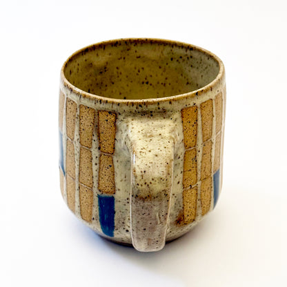 Hand-thrown stoneware mug with a warm speckled glaze and retro geometric grid pattern in tan, deep blue, gray-blue, and muted coral accents. The rounded mug features a curved handle, glossy finish, and mid-century-inspired design. Photographed against a soft white background.
