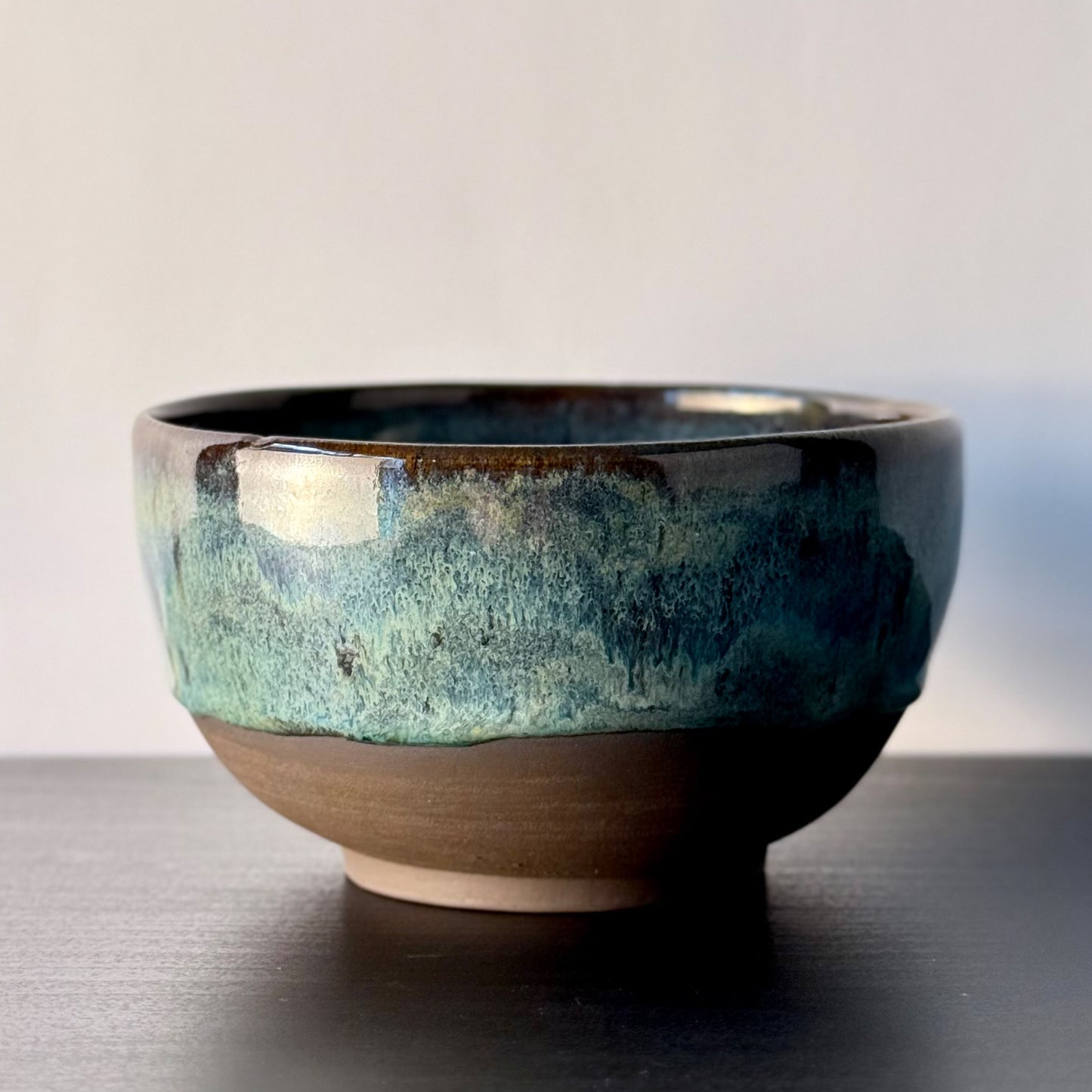 A handmade ceramic bowl with a textured, mottled glaze in shades of teal, blue, and brown, sitting on a dark wooden surface against a neutral light gray background.