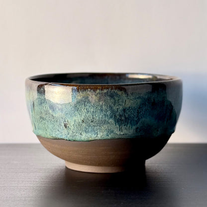 A handmade ceramic bowl with a textured, mottled glaze in shades of teal, blue, and brown, sitting on a dark wooden surface against a neutral light gray background.