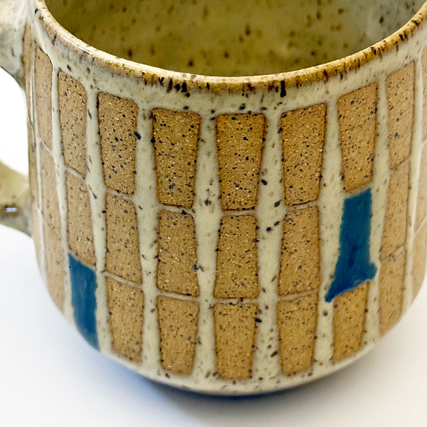 Hand-thrown stoneware mug with a warm speckled glaze and retro geometric grid pattern in tan, deep blue, gray-blue, and muted coral accents. The rounded mug features a curved handle, glossy finish, and mid-century-inspired design. Photographed against a soft white background.
