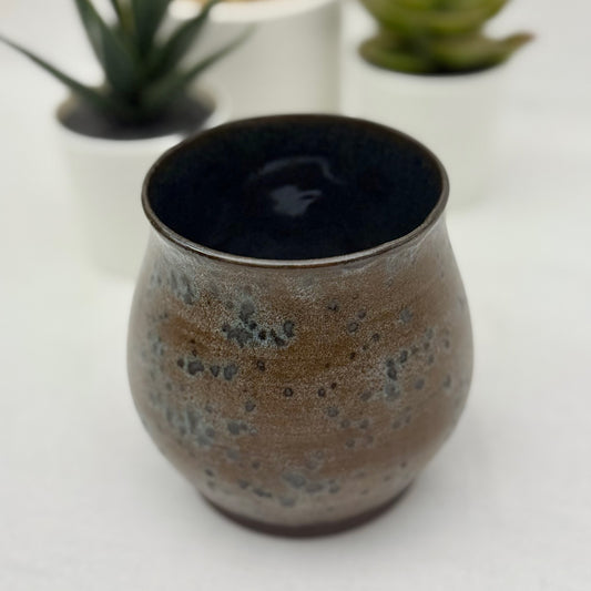 Ceramic cup with speckled design on a white surface with blurred plants in the background