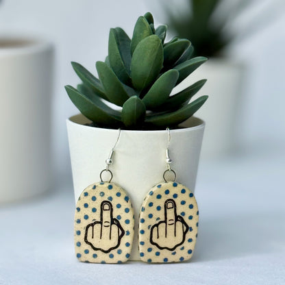 Earrings with middle finger design on polka dot background in front of a potted plant.