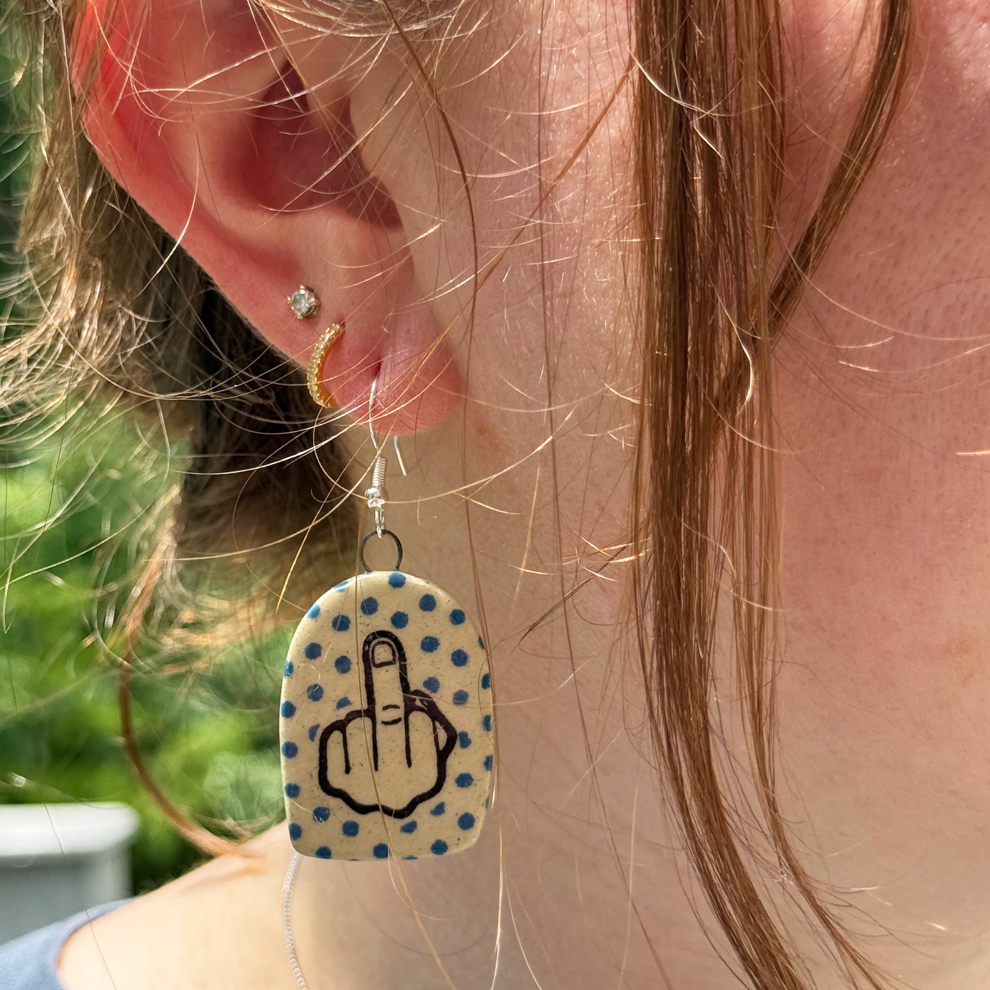 Earring with middle finger symbol on a polka dot background worn by a person.