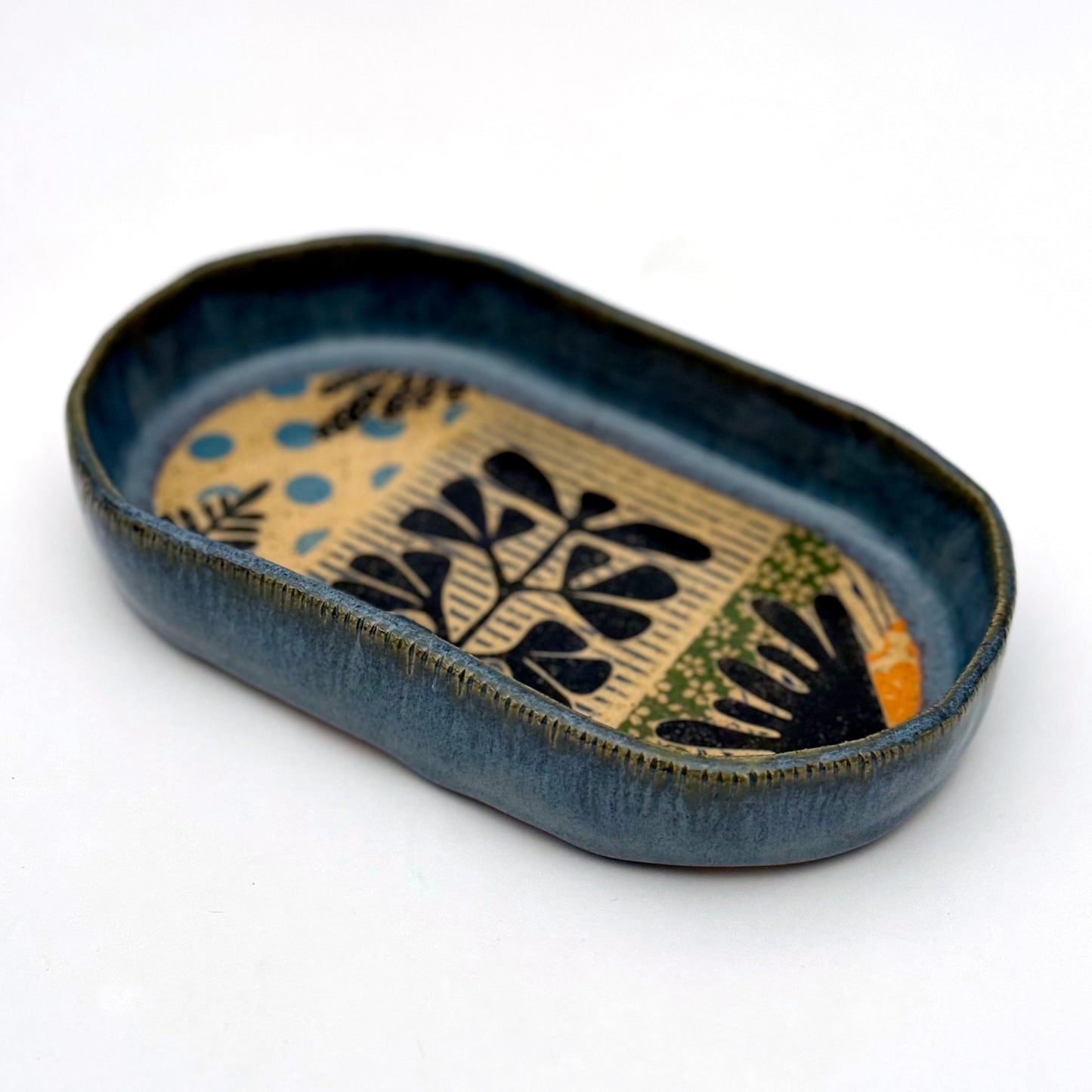 A shallow, oval ceramic trinket tray with a blue exterior rim. The interior floor of the tray features botanical patterns in black, including stylized leaves and stems, set against a speckled buff clay background with pops of orange and light blue polka dots.