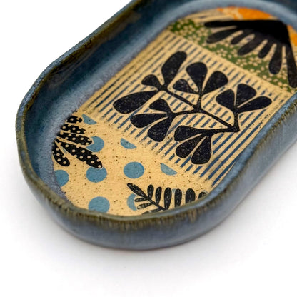 A shallow, oval ceramic trinket tray with a blue exterior rim. The interior floor of the tray features botanical patterns in black, including stylized leaves and stems, set against a speckled buff clay background with pops of orange and light blue polka dots.