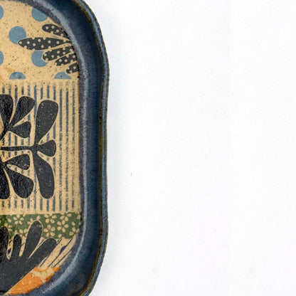 A shallow, oval ceramic trinket tray with a blue exterior rim. The interior floor of the tray features botanical patterns in black, including stylized leaves and stems, set against a speckled buff clay background with pops of orange and light blue polka dots.