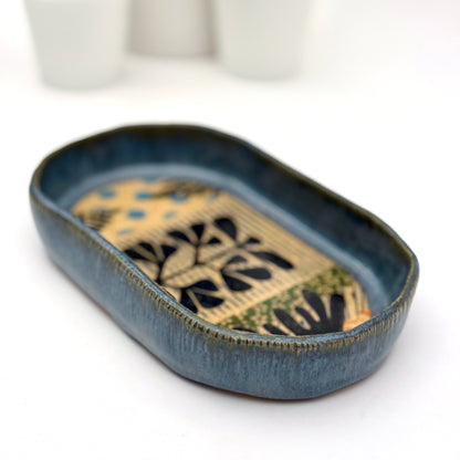 A shallow, oval ceramic trinket tray with a blue exterior rim. The interior floor of the tray features botanical patterns in black, including stylized leaves and stems, set against a speckled buff clay background with pops of orange and light blue polka dots.