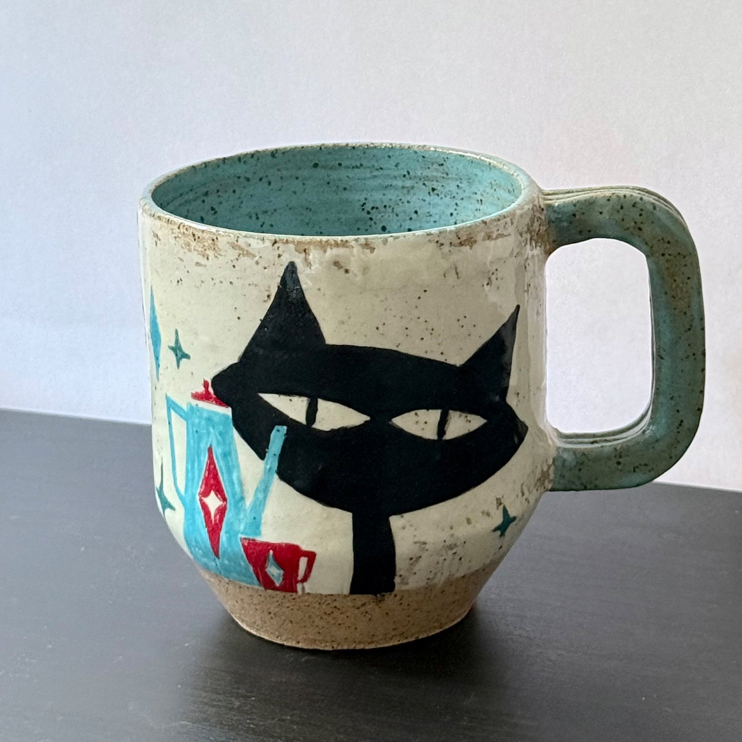 Handmade ceramic mug featuring a black mid-century cat design with turquoise starbursts and red accents on a speckled cream background, with a blue interior and handle.