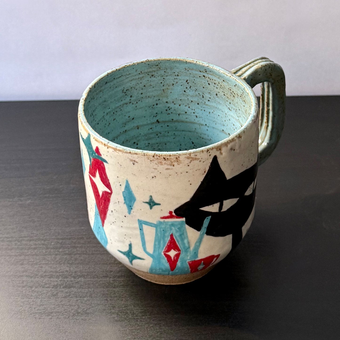 Handmade ceramic mug featuring a black mid-century cat design with turquoise starbursts and red accents on a speckled cream background, with a blue interior and handle.
