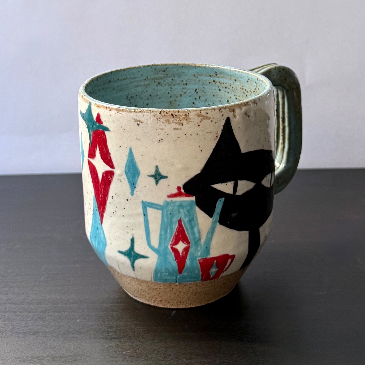 Handmade ceramic mug featuring a black mid-century cat design with turquoise starbursts and red accents on a speckled cream background, with a blue interior and handle.