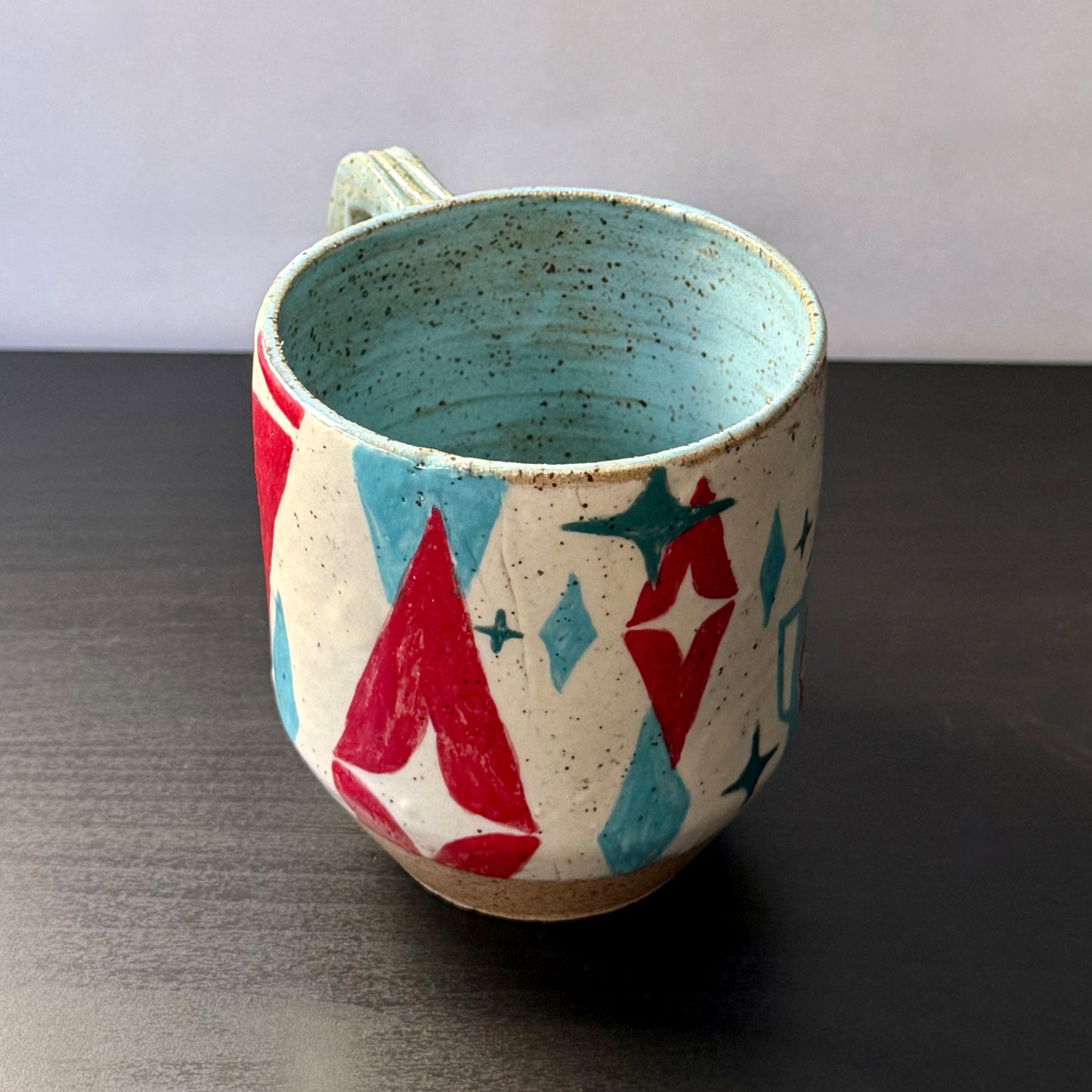 Handmade ceramic mug featuring a black mid-century cat design with turquoise starbursts and red accents on a speckled cream background, with a blue interior and handle.