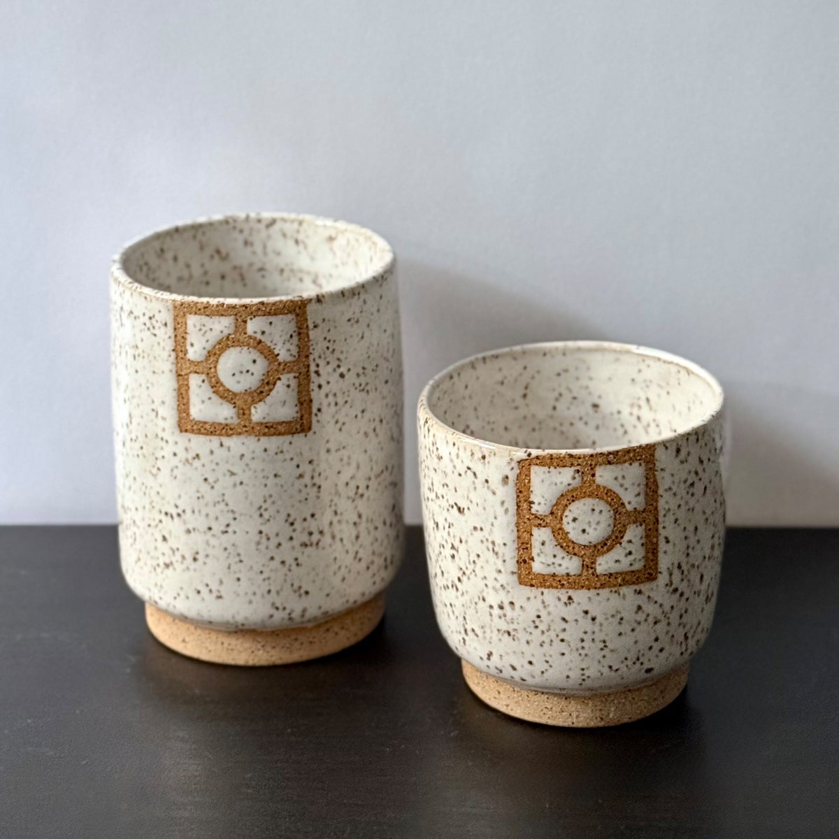 Two handmade ceramic tumblers with a speckled white glaze, exposed stoneware foot rings, and square geometric breeze-block motifs on the front. One tumbler is slightly taller than the other, with both featuring warm natural clay details against the light glaze.
