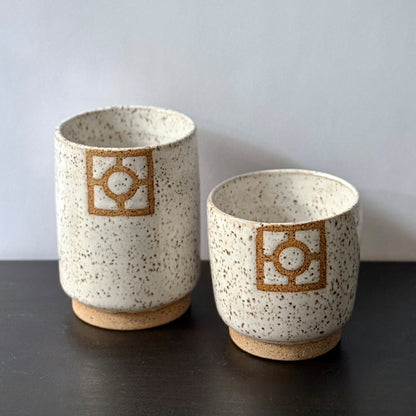 Two handmade ceramic tumblers with a speckled white glaze, exposed stoneware foot rings, and square geometric breeze-block motifs on the front. One tumbler is slightly taller than the other, with both featuring warm natural clay details against the light glaze.